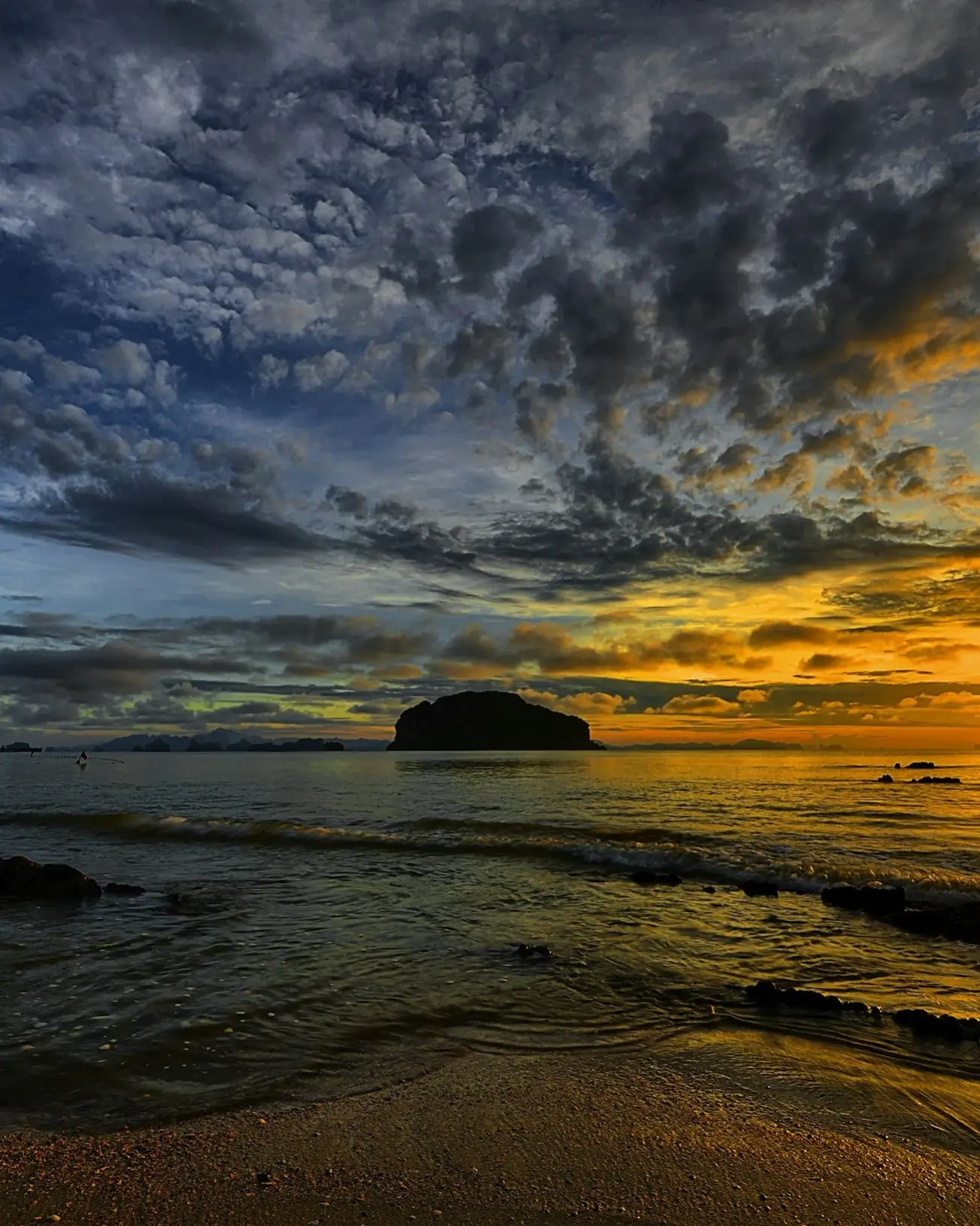 A sunset over the ocean with dark and orange clouds, a rocky shoreline in the foreground and a distant landmass