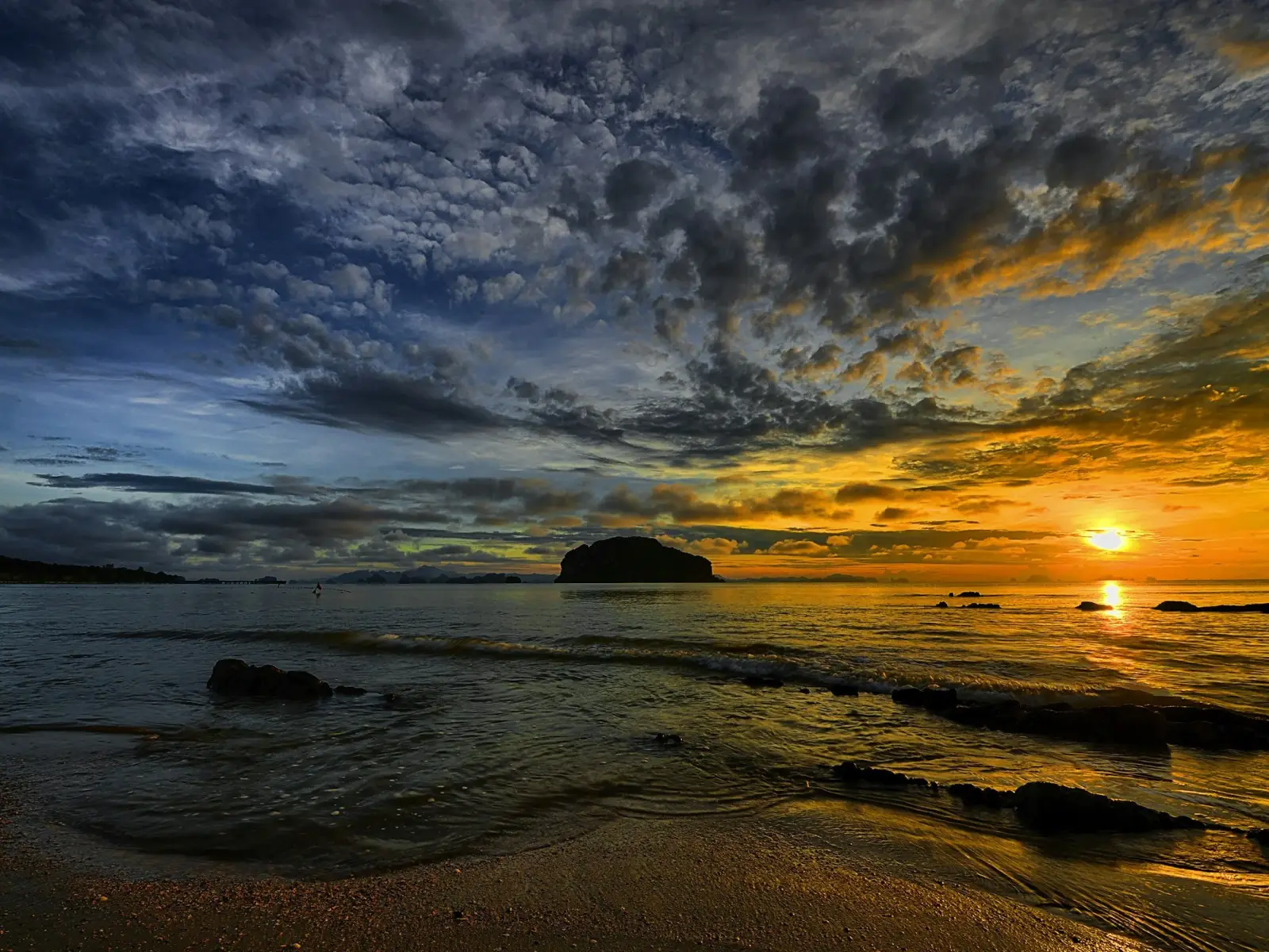 A sunset over the ocean with dark and orange clouds, a rocky shoreline in the foreground and a distant landmass