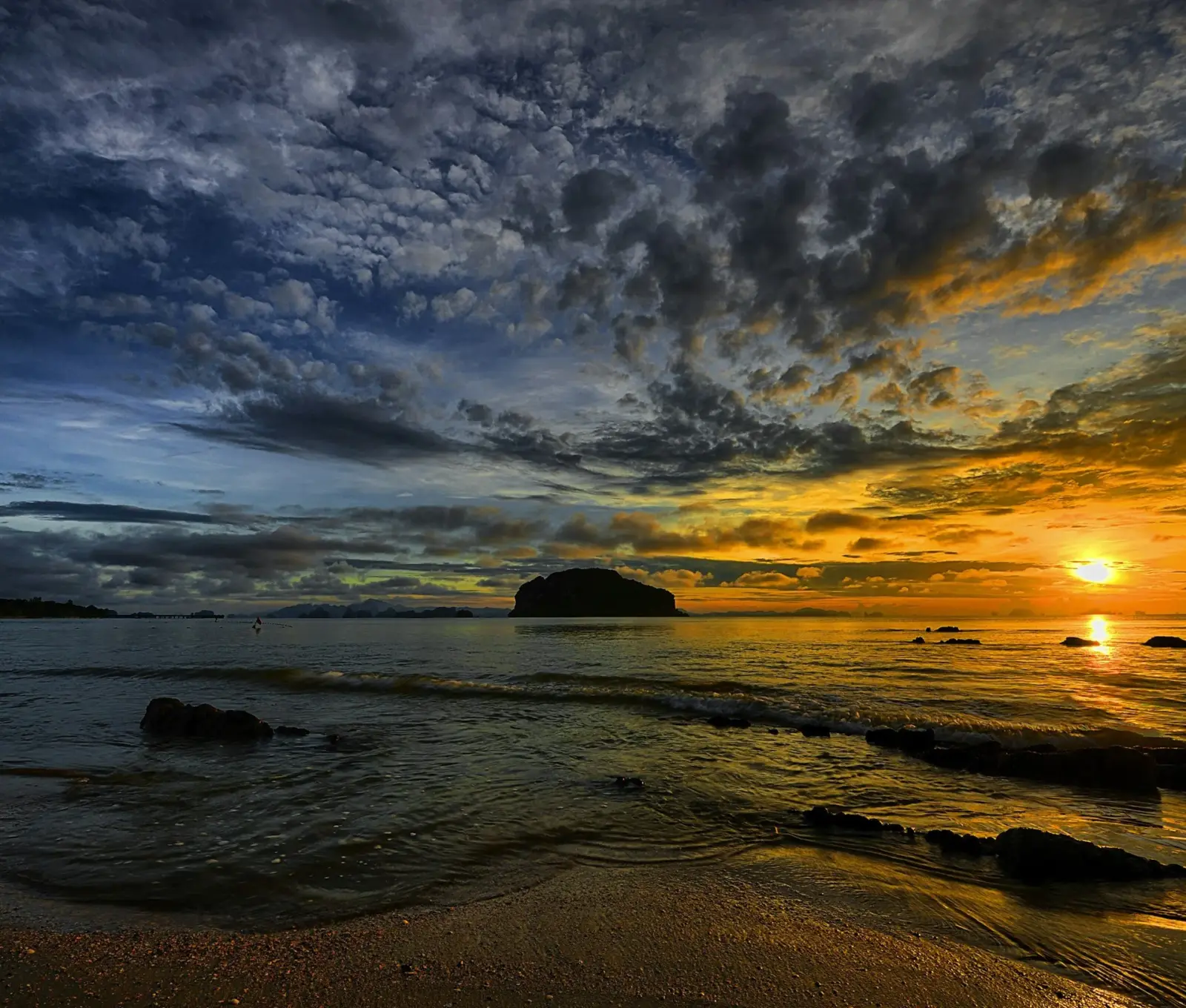 A sunset over the ocean with dark and orange clouds, a rocky shoreline in the foreground and a distant landmass